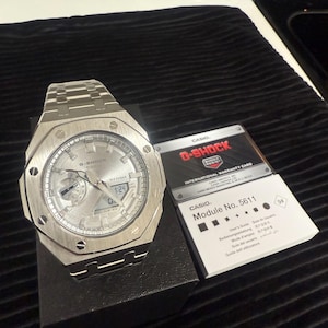 May include: A silver G-Shock watch with a matching metal band, resting on a black surface. The watch face displays the time and date. A Casio warranty card is next to the watch.