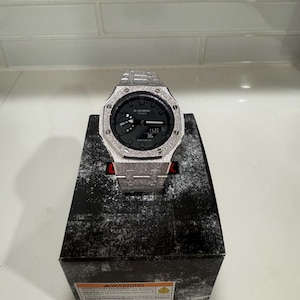 Op de afbeelding: Een zilverkleurig G-Shock horloge met een getextureerde bezel en band, gepresenteerd op een zwarte doos. De wijzerplaat van het horloge is donker met een digitale tijdweergave. Het horloge staat op een zwarte doos met een waarschuwingslabel.