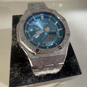 以下が含まれることがあります： ライトブルーの文字盤とデジタル表示を備えた、シルバー調のG-Shock Casioの腕時計。 時計は、テクスチャ加工されたキラキラ光るベゼルとバンドを備えています。 時計は黒い箱に展示されています。