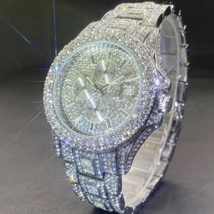 Könnte beinhalten: Eine luxuriöse Armbanduhr mit silbernem Armband und Zifferblatt, vollständig mit glitzernden Kristallen besetzt. Die Uhr verfügt über mehrere Hilfszifferblätter und eine detaillierte Lünette, die ein hochwertiges, glamouröses Design zeigt. Das Armband ist ca. 20 cm lang.
