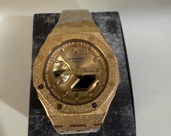 Może przedstawiać: Złoty zegarek G-Shock Casio z teksturowanym, błyszczącym wykończeniem. Zegarek posiada okrągłą tarczę z wieloma tarczami i napisem "G-SHOCK CASIO WR 20BAR". Zegarek jest prezentowany na czarno-szarej, teksturowanej powierzchni.
