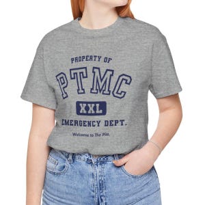 May include: A grey t-shirt with navy blue text that reads "PROPERTY OF PTMC EMERGENCY DEPT." The shirt also has the text "XXL" and "Welcome to The Pitt."