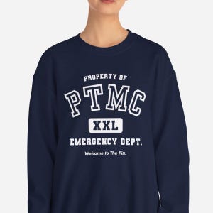 May include: Navy blue sweatshirt with white text that reads "PROPERTY OF PTMC XXL EMERGENCY DEPT. Welcome to The Pitt." The sweatshirt is a crew neck style.