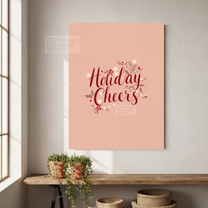 May include: A rectangular canvas print with the words "Holiday Cheers" in a cursive font, surrounded by festive botanical elements and string lights, set against a soft peach background. The print is displayed on a wooden shelf with potted plants.