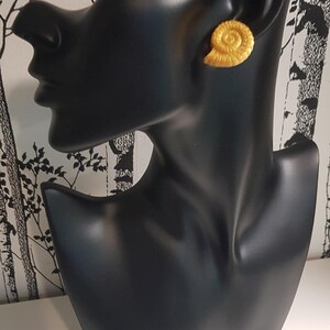 May include: A pair of gold-colored seashell stud earrings. The earrings are displayed on a black mannequin head against a black and white tree-patterned background. The earrings have a spiral design.