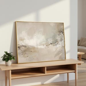 Puede incluir: Impresión artística abstracta en un marco dorado. La obra presenta una paleta de colores neutros con pinceladas beige, blancas y negras. El arte enmarcado se exhibe sobre una consola de madera clara, con una pequeña planta en maceta delante.
