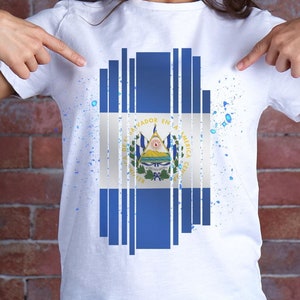 Peut inclure: Un t-shirt blanc avec un motif bleu et blanc du drapeau du Salvador. Le drapeau est composé de bandes verticales avec un blason au centre. Le motif est entouré d'éclaboussures de peinture bleues et blanches.
