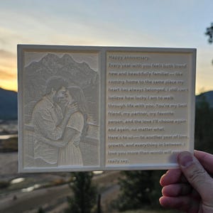 May include: A white, rectangular anniversary plaque with a photo of a couple embracing and a heartfelt message. The plaque is held up against a scenic backdrop of mountains and a river at sunset.