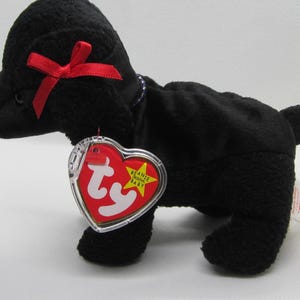 Può includere: Giocattolo a forma di cane in peluche nero con un fiocco rosso e un'etichetta a forma di cuore. L'etichetta recita "Beanie Baby" con una stella gialla. È un oggetto da collezione.