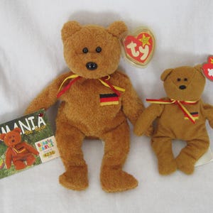 May include: Two brown plush Germania the Bear Beanie Babies toys. The larger bear has a ribbon with the German flag colors and a tag. The smaller bear has a ribbon with the German flag colors and a tag. A tag is attached to each bear.