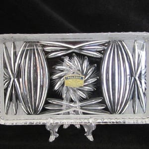 May include: A rectangular, clear glass serving tray with intricate cut glass designs. The tray features a central floral pattern and elongated oval shapes on either side. The edges have a textured, starburst pattern. A small tag reads "POLONIA".