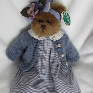 Könnte beinhalten: Ein Teddybär mit einem blauen Vichy-Kleid, einem passenden blauen Strickjäckchen und einem Hut mit Blumen. Der Bär hat ein Etikett mit der Aufschrift "Bearington Collection". Das Outfit ist mit kleinen rosa Rosen verziert.
