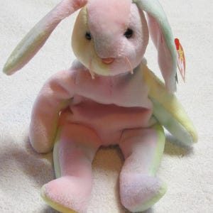 Può includere: Un coniglietto di peluche dai colori pastello. Il coniglio ha lunghe orecchie, un corpo rosa e accenti gialli e verdi. È seduto con le gambe distese. Un'etichetta rossa è attaccata a un orecchio.