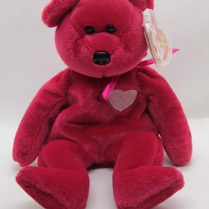 May include: A magenta teddy bear with a heart-shaped patch on its chest. The bear has black eyes and nose, and a pink ribbon. A clear tag is attached to the ear. The plush toy is a vibrant shade of pink.