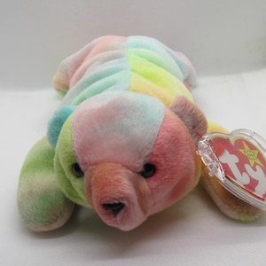 Puede incluir: Un oso de peluche colorido con un patrón de teñido anudado arcoíris. El oso tiene una combinación de colores rosa, verde, amarillo y azul. Tiene ojos negros y una etiqueta roja adjunta a su oreja.