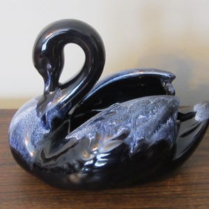 Puede incluir: Figura de cisne de cerámica negra brillante con detalles en azul y blanco en las alas. El cisne tiene un cuello curvado y un área ahuecada, posiblemente para uso decorativo.