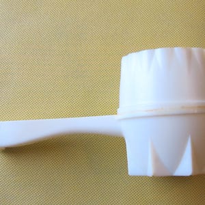 Puede incluir: Un utensilio de cocina de plástico blanco con un mango y una cabeza texturizada en forma de copa. La cabeza tiene una serie de hendiduras triangulares en la base. El mango es curvo.