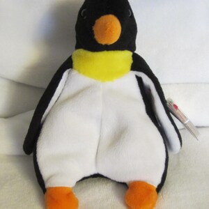 Peut inclure: Une peluche de pingouin avec une tête, un dos et des ailes noirs, un ventre blanc et des pieds et un bec orange. Le pingouin a un col jaune. Une petite étiquette est attachée à l'aile du pingouin.