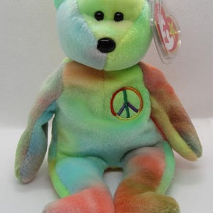 May include: A colorful tie-dye teddy bear plush toy with a peace sign embroidered on its chest. The bear has black eyes and a black nose. The tag is attached to the ear. The bear is a mix of green, blue, orange, and pink.