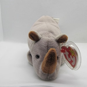 May include: A soft toy rhinoceros in grey and brown. The rhino has small, brown ears and a brown horn. A red and yellow tag is attached to the toy.