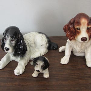 Puede incluir: Tres figuras de perros de cerámica. Una es un perro blanco y negro tumbado, otra es un cachorro pequeño blanco y negro, y la tercera es un perro marrón y blanco sentado erguido. Las figuras están sobre una superficie marrón oscuro.