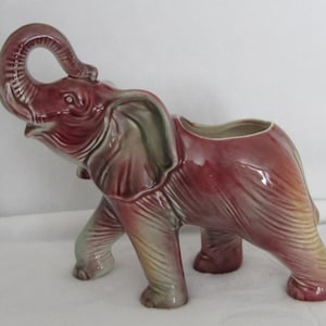 May include: A ceramic elephant planter in shades of burgundy, green, and yellow. The elephant has a raised trunk and a planter opening on its back. The ceramic has a glossy finish and is a decorative item.