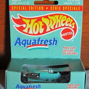 Puede incluir: Un coche de carreras Hot Wheels AquaFresh de edición especial en su embalaje original. La caja es de color verde azulado con el logotipo de Hot Wheels y el nombre de la marca AquaFresh. El embalaje incluye el texto "Race Car" y "Voiture de Course".
