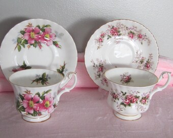 Puede incluir: Dos tazas y platillos blancos con diseños florales. Un juego presenta flores rosas con hojas verdes, el otro tiene rosas rosas. Ambos juegos tienen ribetes dorados y están sobre una superficie rosa.