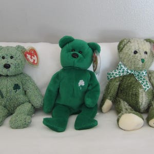 May include: Three plush teddy bears in shades of green. Each bear has a different design, including a shamrock, a bow, and a tag. The bears are sitting on a white surface.