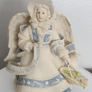 Puede incluir: Figura de ángel color crema con detalles en azul, con sombrero y cesta de flores. El ángel tiene grandes alas y una pancarta que dice "Grandmothers Like You Are Precious And Few."