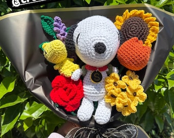 Puede incluir: Un ramo de ganchillo hecho a mano con un personaje de Snoopy blanco con un collar rojo y un colgante dorado. El ramo incluye flores de ganchillo en amarillo, naranja y rojo, envuelto en papel negro y atado con una cinta negra. El texto "@crochetbyyoli" es visible.
