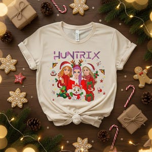 May include: Cream-colored t-shirt featuring the word "HUNTRIX" in purple. The graphic depicts three women in Santa hats and red outfits, surrounded by Christmas elements. A white cat with a red scarf is also present.