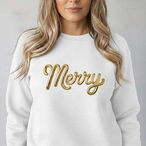 Puede incluir: Sudadera blanca con la palabra "Merry" en letras doradas y cursivas. Las letras doradas tienen un efecto 3D. El fondo es de color gris claro liso.