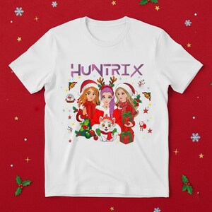 May include: White t-shirt featuring a Christmas-themed graphic with three cartoon women wearing Santa hats and holding gifts. The word "HUNTRIX" is printed across the top in purple. A cat wearing a scarf is also in the graphic.