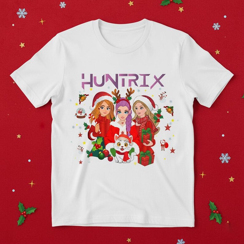 May include: White t-shirt featuring a Christmas-themed graphic with three cartoon women wearing Santa hats and holding gifts. The word "HUNTRIX" is printed across the top in purple. A cat wearing a scarf is also in the graphic.