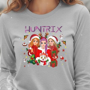 May include: A light gray long-sleeve shirt featuring a graphic of three cartoon women in Christmas attire, with the word "HUNTRIX" above them. The women are surrounded by Christmas decorations and a cat.