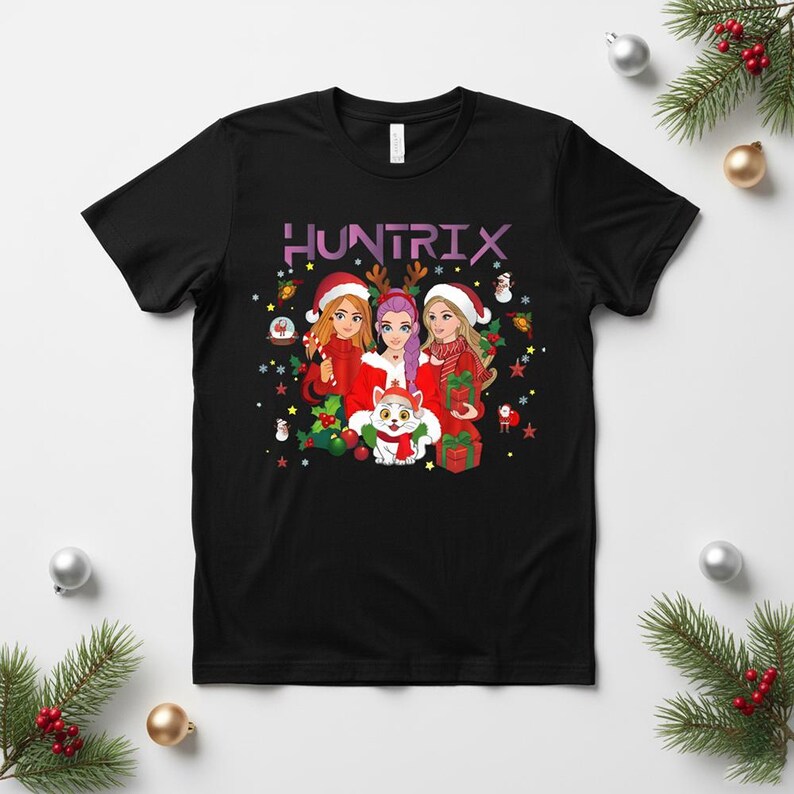 May include: Black t-shirt featuring a festive graphic with the word "HUNTRIX" above a group of three women in Santa hats and a cat wearing a scarf. The design includes Christmas elements like snowflakes, ornaments, and gifts.