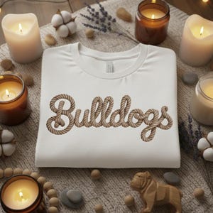 Puede incluir: Una camiseta blanca de cuello redondo con la palabra "Bulldogs" en una fuente de estilo cuerda. La camiseta está rodeada de velas encendidas, capullos de algodón y una figura de bulldog de madera, creando un ambiente acogedor y decorativo.