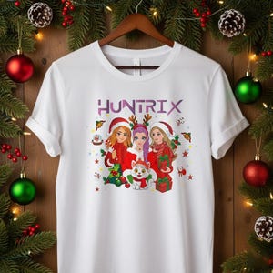 May include: White t-shirt featuring the word "HUNTRIX" in purple letters above a graphic of three women in Santa hats and red outfits, with a cat wearing a scarf. The design includes Christmas elements like stars and gifts.