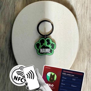 Puede incluir: Una etiqueta verde con forma de pata de mascota con el nombre "NAME" en letras blancas, sujeta a un llavero negro. La etiqueta se muestra en un posavasos circular blanco, con un smartphone y una tableta que muestran información de contacto.