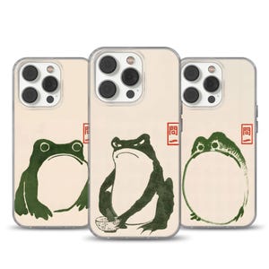 May include: Three phone cases featuring a cream background and green frog illustrations. The frogs are depicted in various poses and expressions. Each case includes a red rectangular stamp with Japanese text.