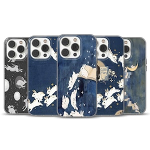 May include: A collection of phone cases with whimsical designs. The cases feature illustrations of rabbits, moons, stars, and open books against dark blue and black backgrounds. The cases are made of clear plastic.