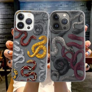 Puede incluir: Dos fundas para teléfono con fondo gris y un patrón de serpientes coloridas. Una funda presenta serpientes rojas, amarillas y blancas, mientras que la otra tiene serpientes rojas, negras y grises. Las fundas están diseñadas para un smartphone.