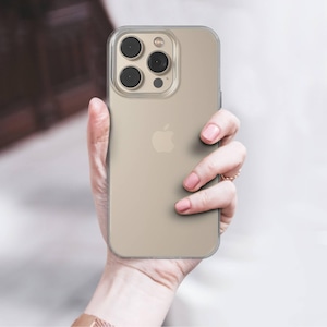 Può includere: Una custodia trasparente per telefono con un telefono color oro all'interno. Il telefono ha una fotocamera a tripla lente e il logo Apple. La custodia è tenuta in mano, con un orologio e un anello d'oro visibili.
