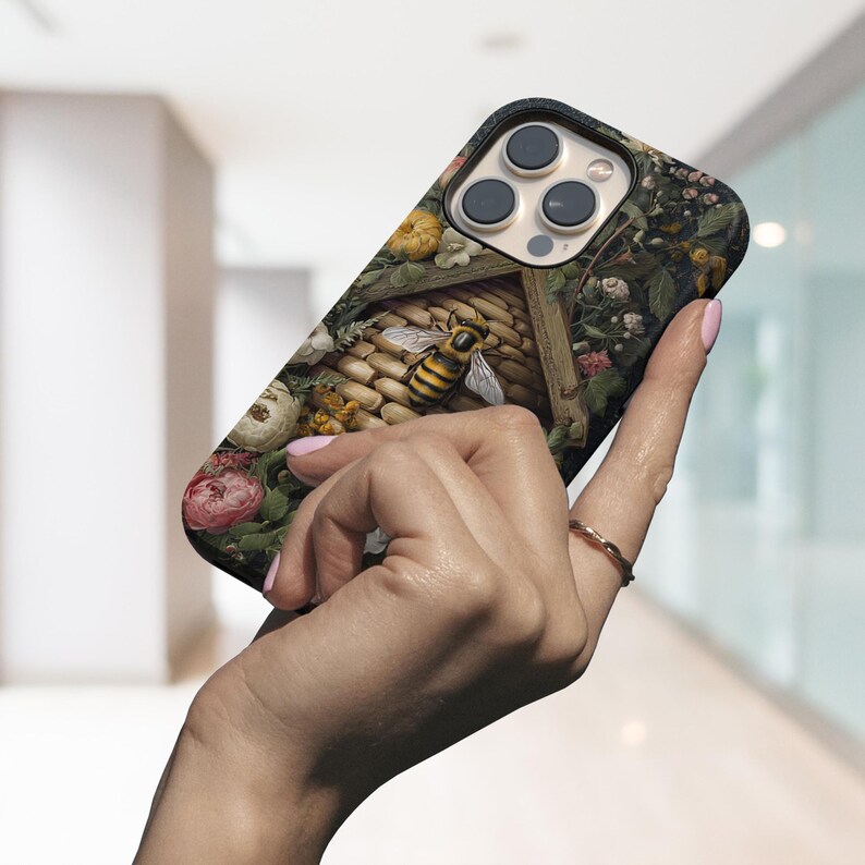 May include: A phone case featuring a detailed illustration of a bee and a beehive surrounded by flowers. The case has a black border and a glossy finish, with a design that incorporates shades of yellow, brown, and green.