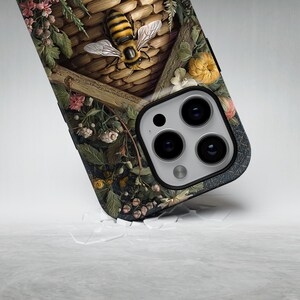 May include: A phone case featuring a detailed illustration of a bee hovering near a woven basket, surrounded by flowers and foliage. The case has a dark background with a textured appearance and a camera cutout.