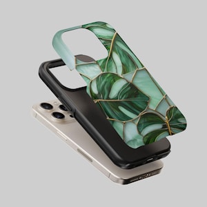 Può includere: Una custodia per telefono con un design a foglie di monstera verdi e oro. La custodia è composta da tre strati: una parte superiore verde chiaro, una parte centrale nera e una base per telefono argentata. Il design delle foglie di monstera presenta sfumature di verde e oro.