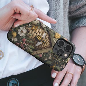 May include: A phone case featuring a detailed illustration of a bee and a honeycomb, surrounded by flowers and foliage. The case has a black camera module and a dark green and brown floral design.