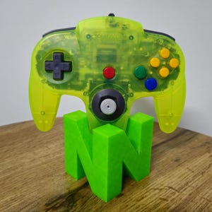 Könnte beinhalten: Ein transparentes neongrünes Nintendo 64-Gamepad mit schwarzen Akzenten und bunten Knöpfen, das auf einem hellgrünen "N64"-Logo ruht. Ein klassisches Gaming-Zubehör.