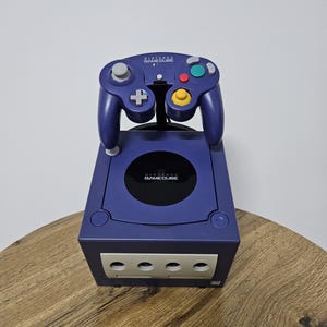 Könnte beinhalten: Eine violette Nintendo GameCube-Konsole mit passendem Controller. Der Controller hat ein Steuerkreuz, farbige Tasten und Analog-Sticks. Die Konsole hat ein Laufwerk und mehrere Anschlüsse. Der GameCube wird auf einer Holzoberfläche präsentiert.